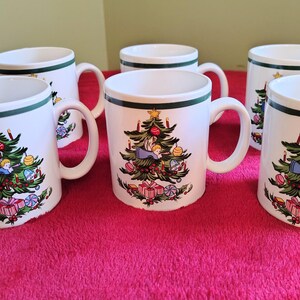 May include: Six white ceramic mugs with a green rim and handle. Each mug features a Christmas tree design with ornaments, gifts, and a star. The mugs are arranged on a pink surface.