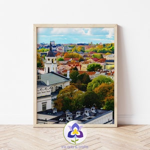 Puede incluir: Una pintura de acuarela de una ciudad con un alto campanario de iglesia en primer plano. La ciudad está pintada en tonos de marrón, rojo y verde, con un cielo azul y nubes blancas en el fondo.