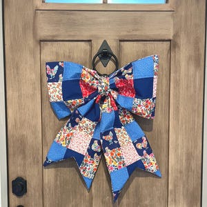 May include: A large, decorative bow made of patchwork fabric, featuring a mix of blue, red, and floral patterns. The bow is attached to a wooden door with a black metal door knocker. The bow's design includes rooster and floral motifs.