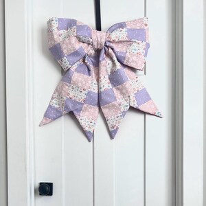 May include: A large, decorative bow made of patchwork fabric in shades of lavender, pink, and white. The bow is attached to a black ribbon and hangs on a white door. The fabric has a floral pattern and the words "HE IS RISEN" are printed on some of the squares.