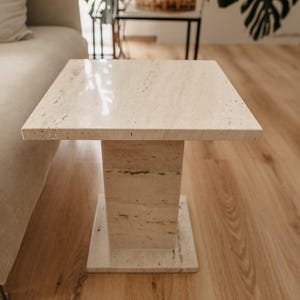Pode incluir: Uma pequena mesa auxiliar quadrada de travertino branco com um tampo liso e polido e uma única perna quadrada grossa.