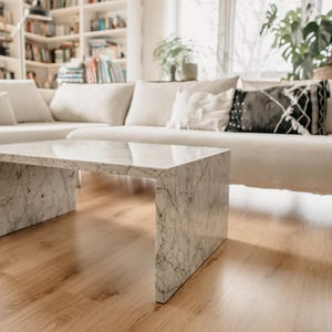 Könnte beinhalten: Ein weißer Marmor-Couchtisch mit einer rechteckigen Tischplatte und einem einzigartigen, offenen Gestelldesign. Der Tisch steht auf einem hellen Holzboden in einem Wohnzimmer.