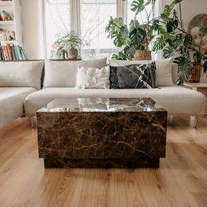 May include: A modern living room featuring a beige sectional sofa, a marble-look coffee table, and hardwood flooring. Decorative pillows in various patterns and colors adorn the sofa. Potted plants and a bookshelf add to the room's decor.