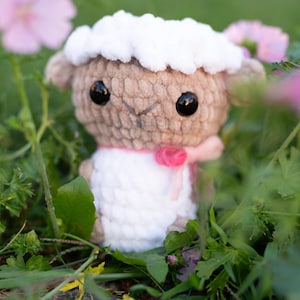 Könnte beinhalten: Ein gehäkeltes Plüschtier-Schaf mit einem weißen, flauschigen Körper, braunem Gesicht und schwarzen Augen. Das Schaf trägt ein rosa Band um den Hals und sitzt in einem Bett aus grünem Gras.
