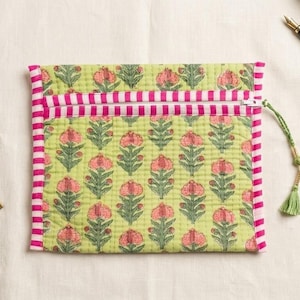 May include: A square quilted pouch featuring a light green fabric with a repeating pink and green floral block print pattern. The pouch has a pink and white striped zipper closure and a green tassel pull. The edges are finished with pink and white striped fabric trim.