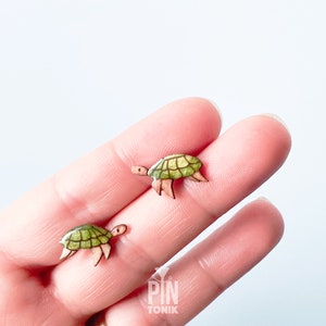 Könnte beinhalten: Zwei kleine Holz-Schildkröten-Figuren, eine grün und eine braun, mit bemalten Details. Die Schildkröten befinden sich auf der Hand einer Person.