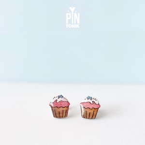 May include: A pair of wooden cupcake earrings with pink icing, white sprinkles, and blue sprinkles. The earrings are made of wood and have a natural finish.