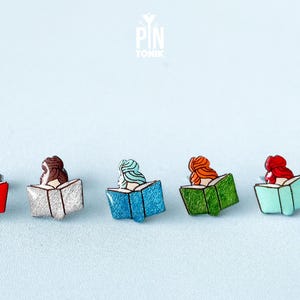May include: Five enamel pins featuring women reading books. The pins are in various colors, including red, silver, blue, green, and turquoise.