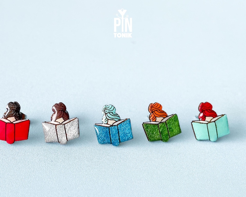 May include: Five enamel pins featuring women reading books. The pins are in various colors, including red, silver, blue, green, and turquoise.