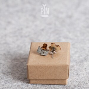 May include: A pair of wooden earrings featuring a person reading a book. The earrings are in a small brown box. The box has the text "PIN TONIK" on the top.