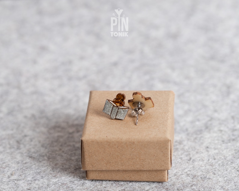 May include: A pair of wooden earrings featuring a person reading a book. The earrings are in a small brown box. The box has the text "PIN TONIK" on the top.