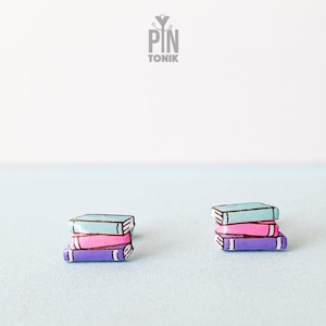 Peut inclure: Une paire de boucles d'oreilles représentant une pile de trois livres. Les livres sont peints dans des tons de bleu, rose et violet.
