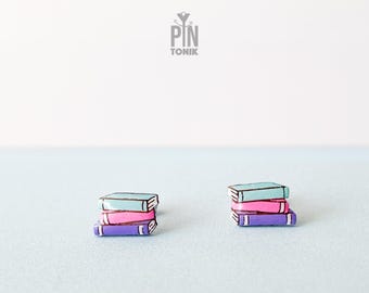 May include: A pair of earrings featuring a stack of three books. The books are painted in shades of blue, pink, and purple.
