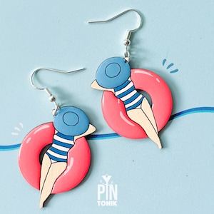 May include: A pair of earrings featuring a pink inflatable ring with a person wearing a blue hat and striped swimsuit floating on top. The earrings are made of wood and have silver hooks.