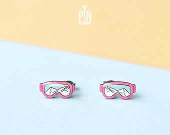 May include: A pair of pink enamel earrings shaped like ski goggles with a white mountain scene inside.