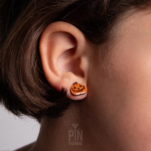 Può includere: Orecchini a bottone a forma di zucca in legno. Gli orecchini sono arancioni con un design a volto intagliato. Gli orecchini sono sull'orecchio di una persona. Un accessorio a tema Halloween.