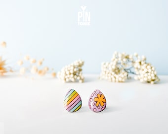 Könnte beinhalten: Ein Paar bunte Ohrringe in Form von Ostereiern. Ein Ohrring hat ein gestreiftes Design in Pink, Gelb und Blau. Der andere Ohrring hat einen rosa Hintergrund mit weißen Punkten und einer gelben Blume.