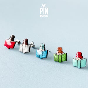 May include: Five pairs of stud earrings featuring a woman reading a book. The books are in different colors: red, white, blue, green, and turquoise.