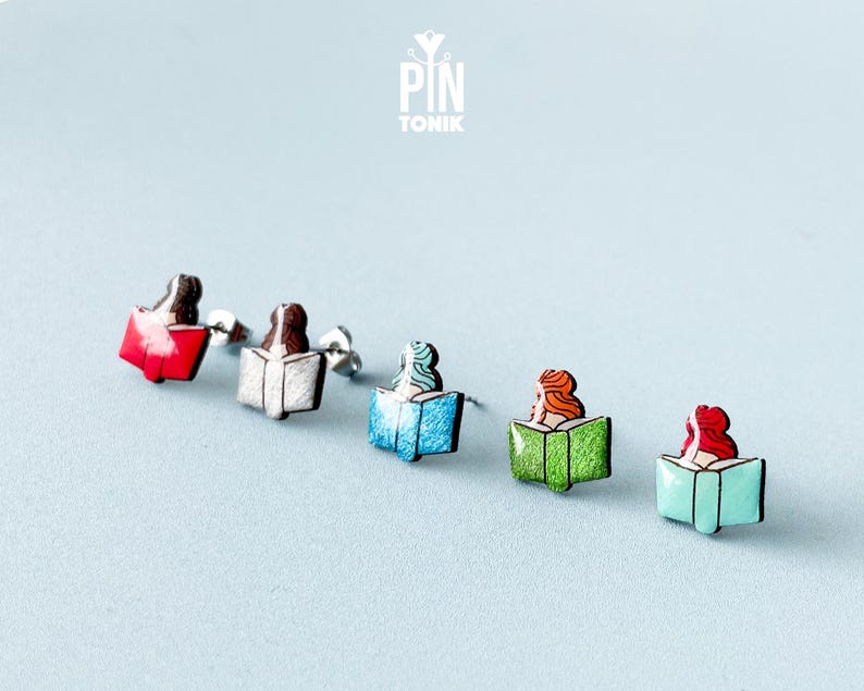May include: Five pairs of stud earrings featuring a woman reading a book. The books are in different colors: red, white, blue, green, and turquoise.