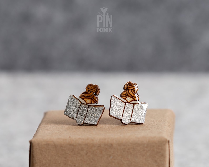 May include: A pair of silver glitter book earrings with a wooden silhouette of a person reading. The earrings are on a brown surface.