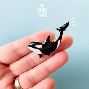 Puede incluir: Un pin de orca en blanco y negro con un acabado brillante. El pin presenta un diseño detallado del cuerpo de la orca, incluyendo su aleta dorsal y cola. El pin se sostiene en una mano sobre un fondo azul claro. El texto "PIN TONIK" está en la parte superior.