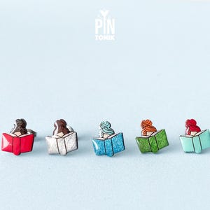May include: Five enamel pins featuring a person reading a book. The pins are in different colors: red, white, blue, green, and turquoise.