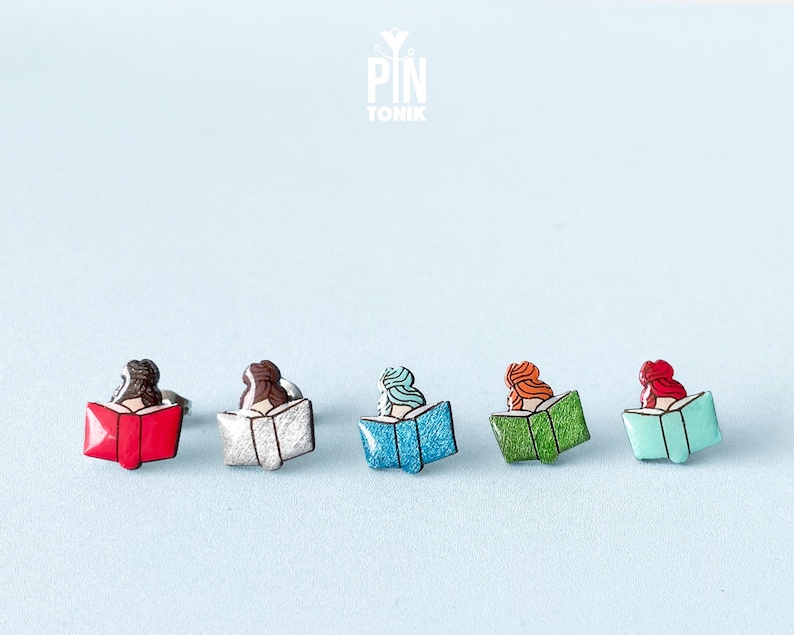 May include: Five enamel pins featuring a person reading a book. The pins are in different colors: red, white, blue, green, and turquoise.