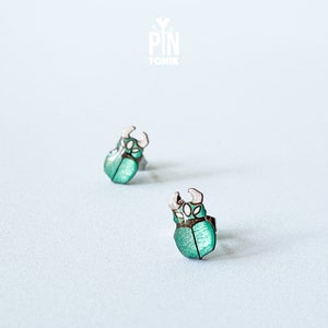 Puede incluir: Un par de pendientes de botón plateados con un diseño de escarabajo verde. Los escarabajos tienen detalles blancos y están colocados sobre un fondo blanco.