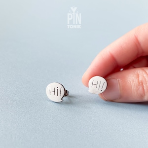 Puede incluir: Un par de pendientes de plata con forma de bocadillo de diálogo con la palabra "Hi!" escrita dentro de cada bocadillo.