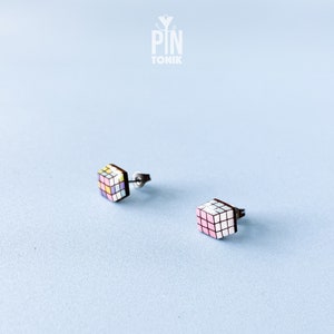 Puede incluir: Pendientes de botón de madera con diseño de Cubo de Rubik. Los cubos tienen un patrón geométrico colorido con tonos pastel de rosa, amarillo, azul y blanco. Los pendientes están sobre un fondo azul claro.