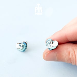 Puede incluir: Un par de pendientes en forma de corazón con un diseño de olas azul y blanco. Los pendientes están hechos de metal y tienen un cierre de poste.
