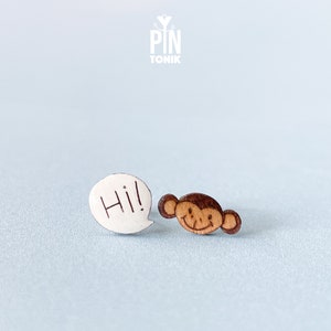 Op de afbeelding: Een paar houten oorbellen met een cartoon-apenkop en een spraakballon met het woord "Hi!" in witte letters.