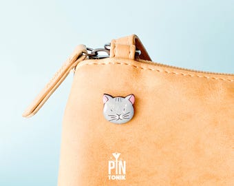 May include: A light brown leather zippered pouch with a gray cat face pin attached. The pin has pink ears and a white nose. The pouch has the text "PIN TONIK" printed on the bottom.