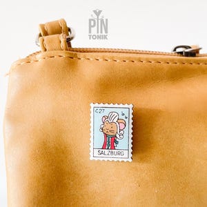 Può includere: Spilla rettangolare in smalto raffigurante un topo dei cartoni animati vestito con un abito rosso e verde, simile a un francobollo. La spilla è fissata a una borsa in pelle marrone chiaro. Il testo "SALZBURG" è visibile.