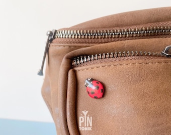 May include: A brown leather fanny pack with a silver zipper and a red ladybug pin attached to the front. The pin has black spots and a silver outline.