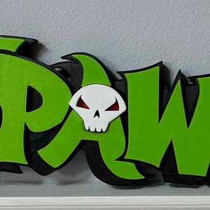 May include: A green and black 3D printed sign that reads "SPAWN" with a white skull with red eyes in the centre.