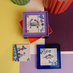 Könnte beinhalten: Drei quadratische Keramik-Untersetzer mit blauen und weißen Blumenmustern. Ein Untersetzer zeigt eine Krabbe, ein anderer zwei Fische und der dritte einen einzelnen Fisch. Die Untersetzer sind in Lila, Orange und Blau gerahmt.