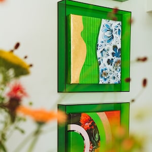 Peut inclure: Deux peintures abstraites encadrées avec des cadres verts. La peinture du haut présente un motif abstrait or et vert avec un motif floral bleu et blanc sur le côté droit. La peinture du bas présente un motif abstrait vert, rose et orange avec un motif floral.