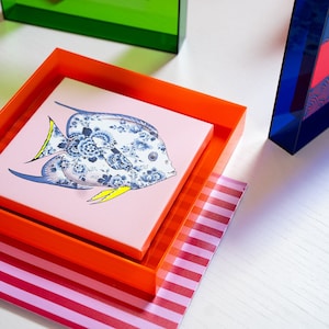 May include: A colorful, framed print of a blue and white fish with floral details on a pink background. The print is displayed in a bright orange frame, which is sitting on a pink and white striped surface.