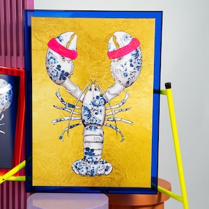 May include: A blue and white floral patterned lobster with pink bands around its claws, set against a gold background in a blue frame.