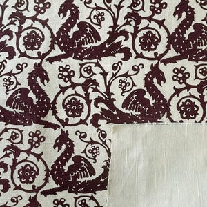 May include: Cream-colored fabric with a repeating pattern of maroon mythical creatures, flowers, and swirling vines. The design is reminiscent of medieval tapestries. A section of plain cream fabric is visible.