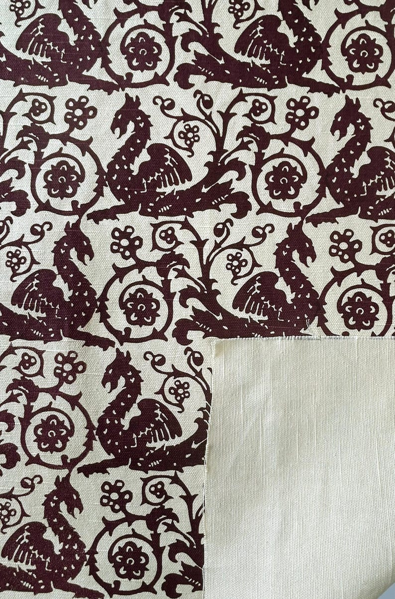 May include: Cream-colored fabric with a repeating pattern of maroon mythical creatures, flowers, and swirling vines. The design is reminiscent of medieval tapestries. A section of plain cream fabric is visible.