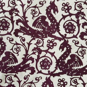 May include: Cream-colored fabric with a repeating pattern of stylized dragons, floral elements, and swirling vines in a deep burgundy color. The design has a vintage or medieval aesthetic, suitable for upholstery or decorative projects.