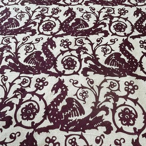 May include: Close-up of a fabric with a repeating pattern of stylized mythical creatures and floral elements in a deep burgundy color against a cream background. The design has a vintage, ornate aesthetic.