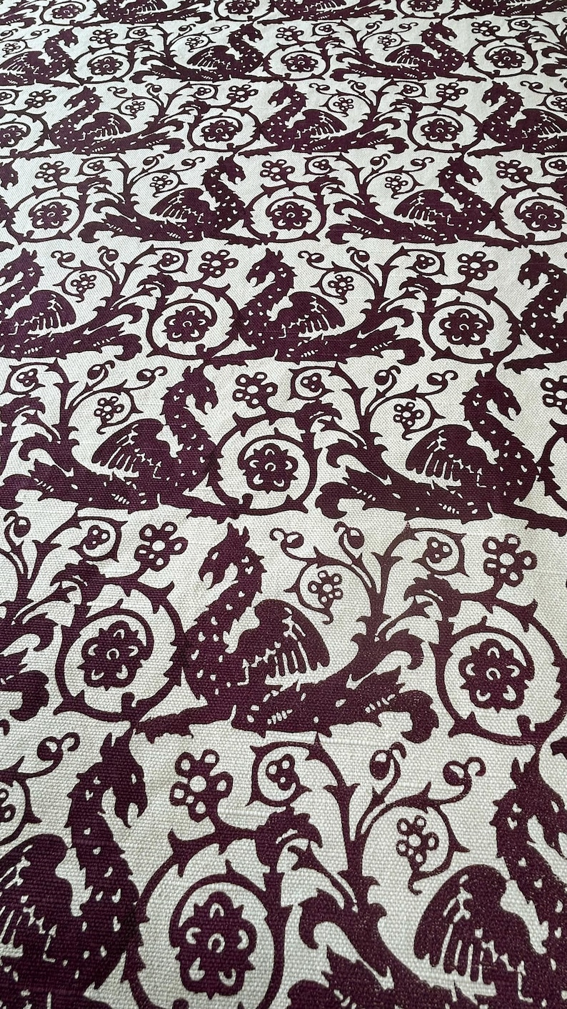 May include: Close-up of a fabric with a repeating pattern of stylized mythical creatures and floral elements in a deep burgundy color against a cream background. The design has a vintage, ornate aesthetic.