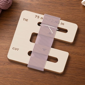 May include: A light-coloured wooden yarn tassel maker with slots and the printed text TIE and CUT. The tool is wrapped with mauve yarn, and the text 75 m in is visible at the top. The device features rectangular cutouts for yarn management.