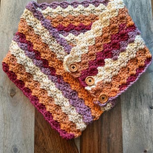 May include: A handmade crochet cowl in warm tones of cream, orange, purple, and burgundy. The cowl features a button closure with three wooden buttons. The textured stitch pattern adds visual interest to this cozy neck warmer.