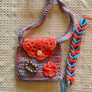 May include: A crocheted purse with a blue and orange color scheme. The purse has a flower and a beaded bracelet attached to it. The purse has a long strap that can be worn over the shoulder.