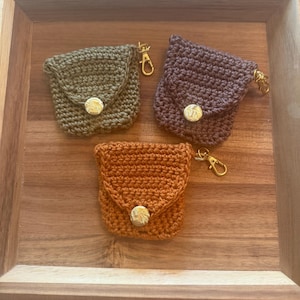 May include: Three small, crocheted pouches in olive green, plum, and burnt orange, each with a gold-colored clasp and button closure. The pouches are displayed in a wooden tray, showcasing their handmade texture and design.