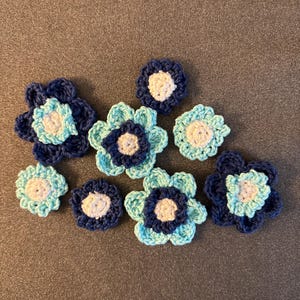 May include: A collection of handmade crochet flowers in various shades of blue and white. The flowers have a layered petal design with a contrasting center. These decorative flowers are ideal for crafting projects.